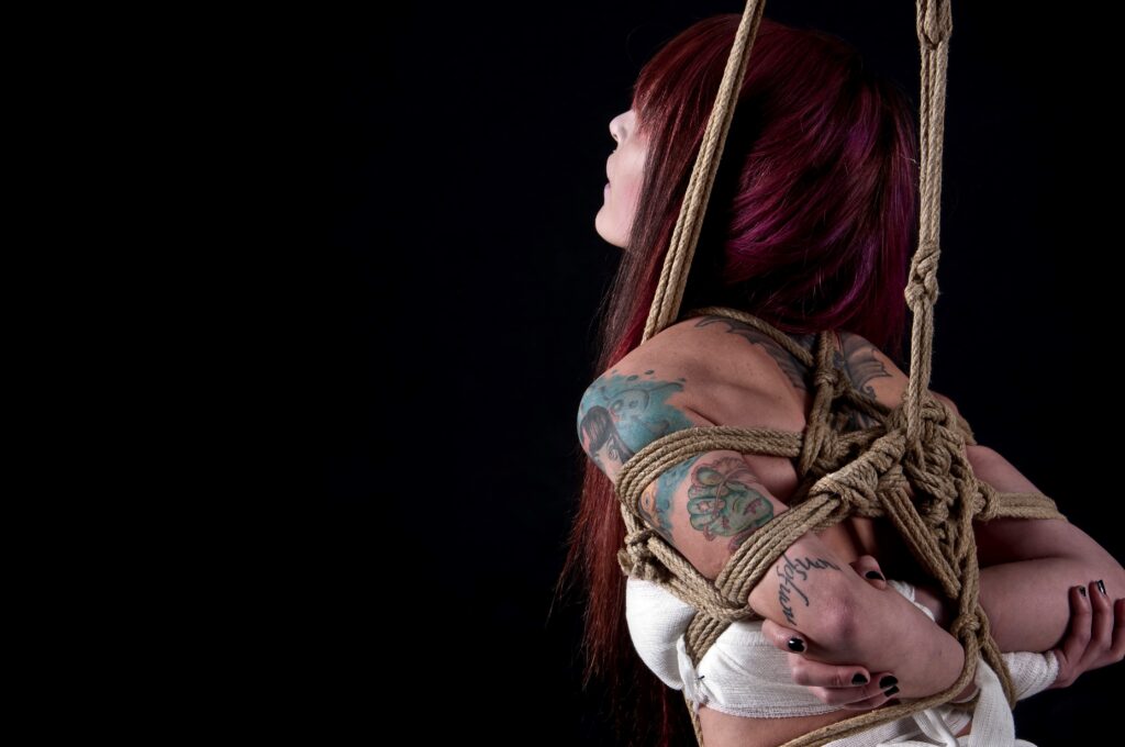 BDSM Escort demonstriert Shibari mit komplexen Seilbindungen auf einer Frau mit roten Haaren und Tattoos, hervorgehoben durch das Spiel von Licht und Schatten.