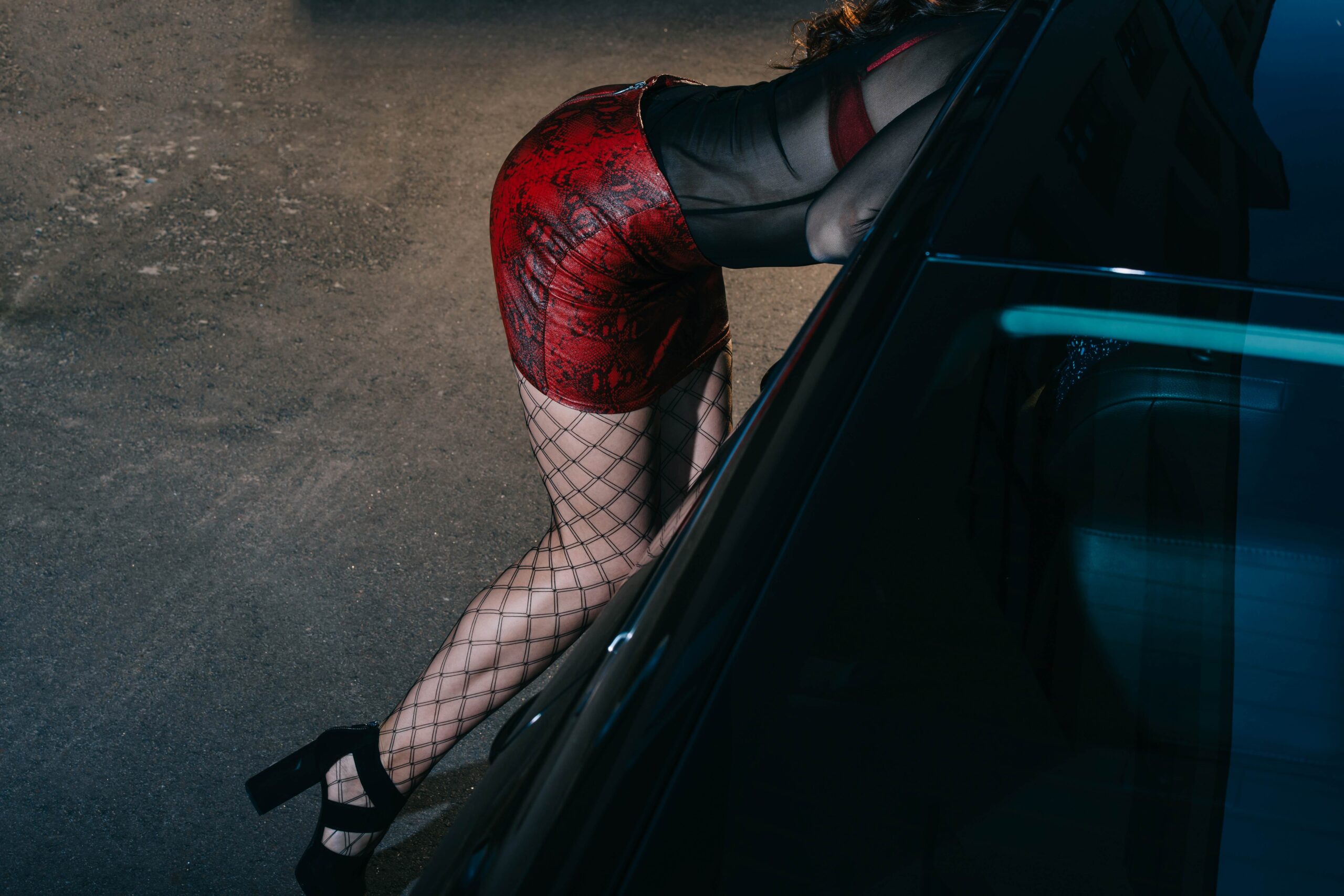 BDSM Escort in verführerischer Pose neben einem dunklen Auto, trägt ein rotes Outfit und Netzstrümpfe, unterstreicht die Kombination aus Eleganz und Provokation in einem urbanen Umfeld